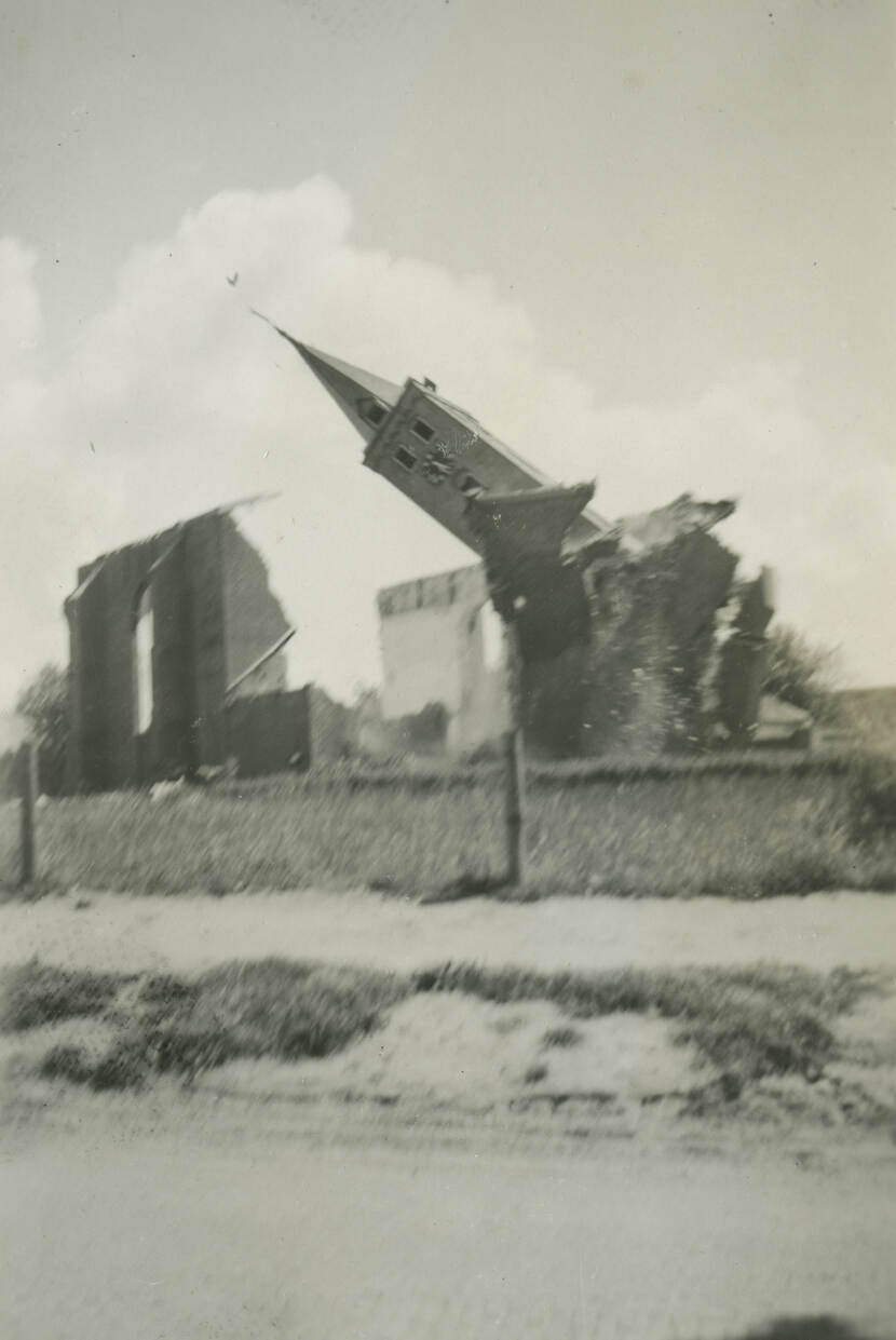 Zwart-witfoto. Een kerktoren wordt gesloopt, de punt valt naar links, terwijl stenen en puin rondvliegen. Links ervan staat nog één kerkmuur overeind. In de voorgrond een weiland en een zandweggetje.