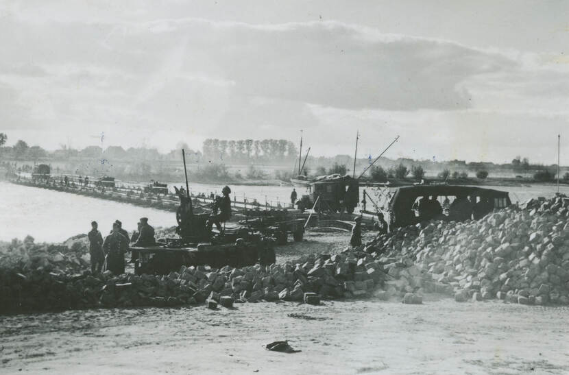 Zwart-witfoto. Militaire voertuigen rijden over een geïmproviseerde brug over een rivier, op de kant staan twee voertuigen op hun beurt te wachten. Zowel op, in en om de wagens staan meerdere militairen. In de voorgrond een grote stapel stenen, in de achtergrond polderlandschap met bomen, op de rivier ligt een bootje.