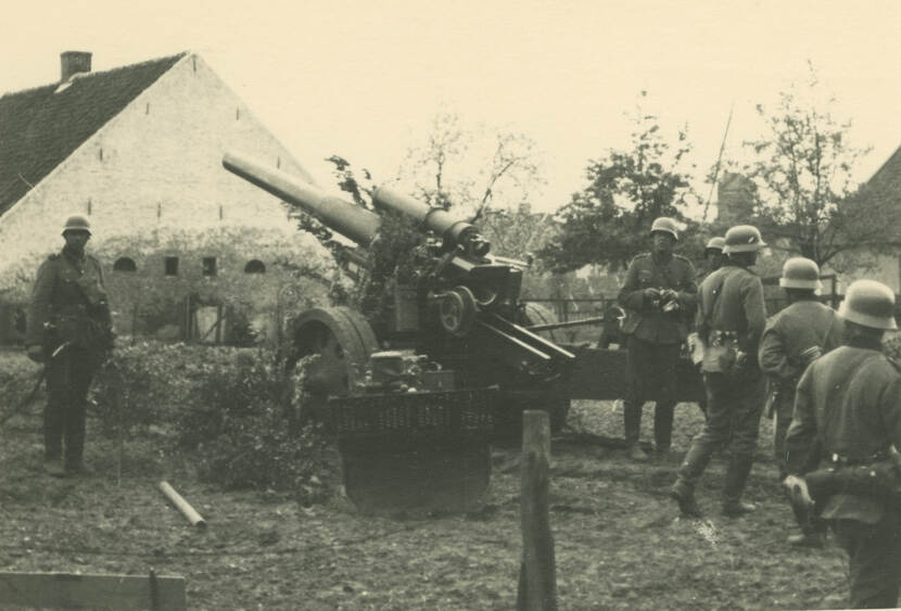 Zwart-witfoto. Een kanon staat in de tuin van een boerderij, daaromheen staan en lopen zes Duitse militairen. Links in de achtergrond een hoge schuur, verder bomen en in de verte een silo.