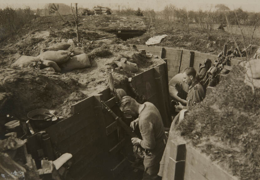 Zwart-witfoto. Een zigzaggende loopgraaf, daarin staan militairen die met hun wapens bezig zijn, de wanden zijn met houten planken bedekt, bovenop liggen zandzakken. In de omgeving polderlandschap met veel jonge boompjes.
