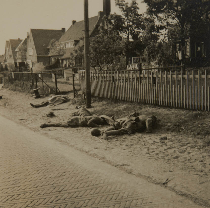 Zwart-witfoto. Vier dode militairen liggen langs de kant van de weg langs een tuinhek, op het hek staat een bordje met de woorden ‘Rust Wat’. In de achtergrond bomen en rijtjeshuizen.