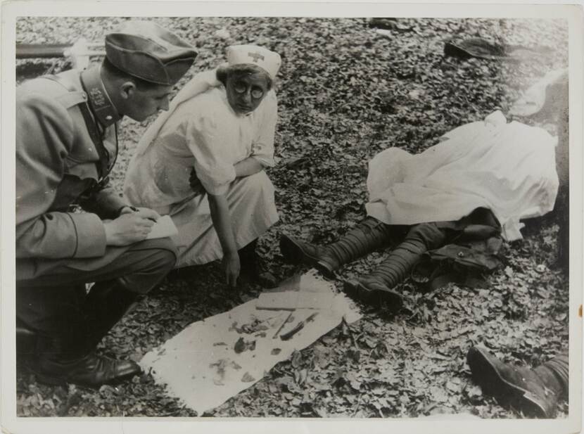 Zwart-witfoto. Een militair en een in het wit geklede verpleegster zitten op hun hurken op de grond, de militair houdt een pen en blocnote op zijn knie. Voor hen ligt een wit doek met attributen, rechts een lichaam dat met een wit deken bedekt is, de benen steken er onderuit.