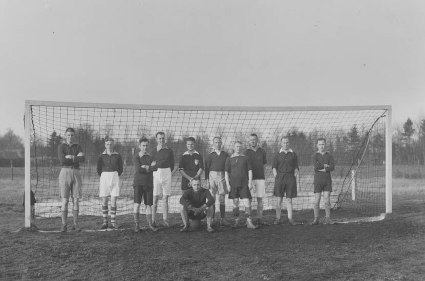 Zwart-witfoto. Tien mannen staan in een groot voetbaldoel, zijn dragen allemaal een voetbaltenue met korte broek en hoge kousen. Vijf dragen een witte broek, vijf een donkere broek, er is weinig eenheid in de kousen. Twee staan met hun armen over elkaar, één met de handen in zijn zakken. Voor hen zit een keeper op een bal. In de achtergrond grasveld, akkerland, hekjes en bomen.