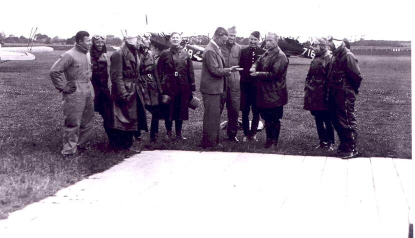 Zwart-witfoto. Een groep van elf mannen staat in een grasveld met elkaar te praten en te lachen, de meesten dragen lange leren jassen, een vliegermuts en vliegerbril op hun hoofd. Een burger in het midden houdt zijn hand op en kijkt ernaar. Op de voorgrond een groot houten terras, in de achtergrond vliegtuigen die aan de grond staan.
