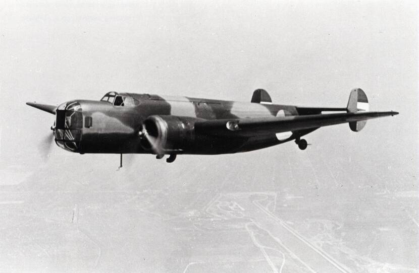 Zwart-witfoto. Een militair vliegtuig vliegt door de lucht, de propeller op de vleugel draait, het heeft een grote, glazen neus. Het toestel is in camouflagekleuren geschilderd, op de zijkant is de roundel van de Luchtmacht aangebracht, de staart is in de Nederlandse driekleur.