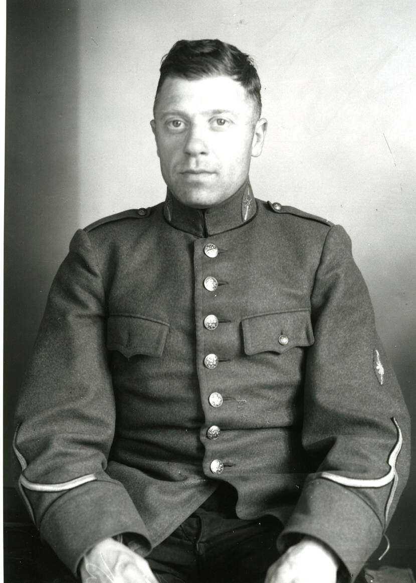 Zwart-witfoto. Portret van een militair, hij kijkt recht in de camera. Hij draagt een uniform met zeven grote knopen op de voorkant en sierlijke onderscheidingstekens aan het uiteinde van de mouwen. Zijn handen houdt hij op zijn bovenbenen.