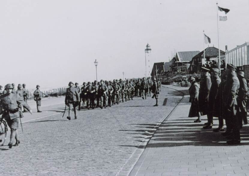 Zwart-witfoto. Militairen marcheren in gelid over een boulevard, rechts staat een groep militairen te kijken, waaronder een oudere vrouw die vooraan staat. In de achtergrond wapperen vlaggen, huizen en een vuurtoren.