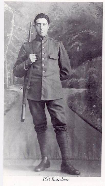Zwart-witfoto. Portretfoto van een militair, hij staat in een fotostudio voor een achtergrond met bomen en een paadje. Hij is geheel te zien, hij draagt een uniform met zwarte kisten. Met zijn rechterhand houdt hij de riem van zijn geweer vast die over zijn schouder hangt, zijn andere hand heeft hij op de rug.