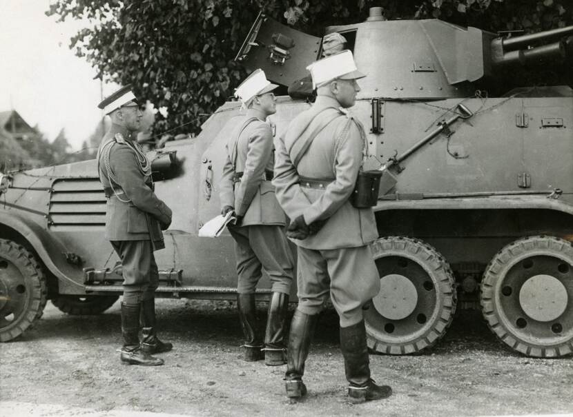 Zwart-witfoto. Drie hoge militairen staan naast een pantserwagen, zij dragen alle drie een witte band om hun pet, de linker heeft meerdere koorden om zijn rechterschouder. De middelste militair houdt papieren in zijn handen, die hij achter zijn rug heeft, ook de rechter heeft de handen op de rug. Vanuit de pantserwagen steekt een vierde militair met zijn hoofd en een arm naar buiten door het openstaande luik. In de achtergrond een boom en linksachter het dak van een huis.