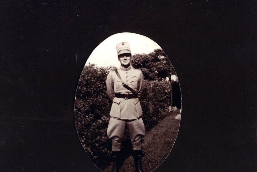 Zwart-witfoto. Een militair staat in een tuin, met zijn handen op de rug. Hij draagt een uniform met een brede riem en een dunnere leren riem schuin over de borst, de klep van zijn pet werpt een schaduw over zijn ogen. In de achtergrond een heg, gras en bomen.