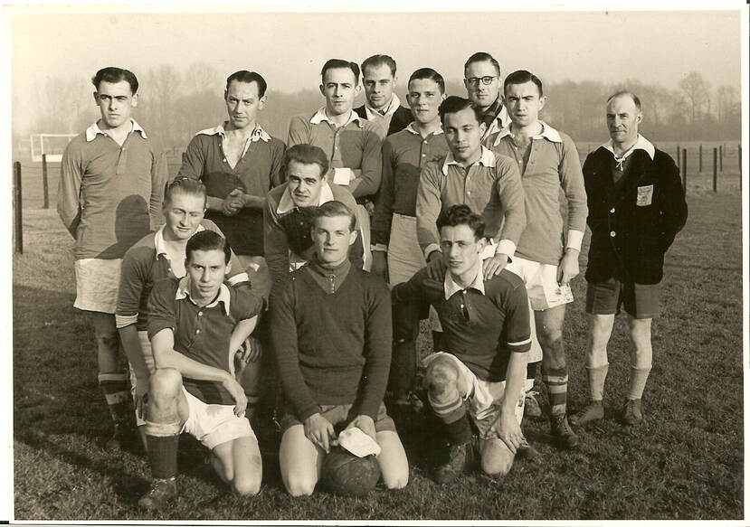 Zwart-witfoto. Groepsfoto van veertien mannen, het merendeel draagt een voetbaltenue met witte broekjes en witte kragen. De voorste drie zitten op hun knieën op de grond, daarachter staan drie voorovergebogen,  de laatste rij staat recht overeind. De man in het midden op de eerste rij heeft een bal tussen zijn benen.