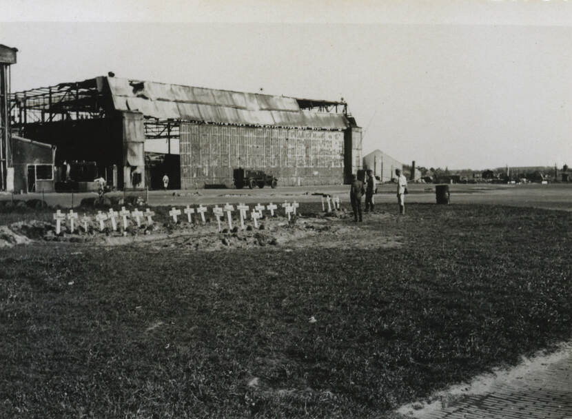 Zwart-witfoto. Een grasveldje met daarachter een grote hangar. De hangar is beschadigd, grote delen van het dak ontbreken. In het grasveldje zijn 27 witte kruisen aangebracht, daarvoor is duidelijk aarde en grond omgewoeld, rechts staan drie mannen naar de kruisen te kijken.