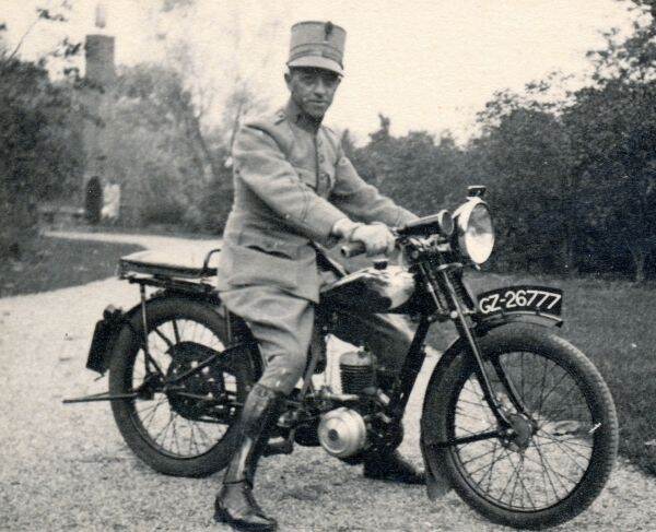 Zwart-witfoto. Een militair zit in uniform op een motorfiets, hij draagt een hoge pet, zwarte laarzen en witte handschoenen. De motorfiets staat op een grindpad, op het spatbord op het voorwiel staat GZ-26777. In de achtergrond struiken, bomen en een toren.