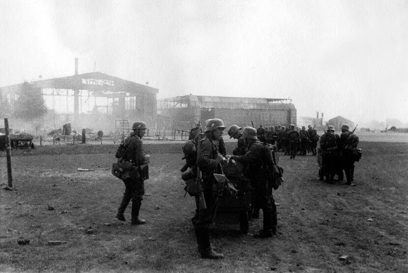 Zwart-witfoto. In de voorgrond staan vijf Duitse militairen in een groepje bij elkaar, rechts lopen twee andere militairen weg van de camera. Daarachter loopt een grotere groep van minimaal tien Duitse militairen in formatie ook weg van de camera. Alle militairen zijn zwaar bepakt en dragen helmen en geweren. In de achtergrond staan twee grote hangars, beide zijn ernstig beschadigd en lijken in brand te staan.