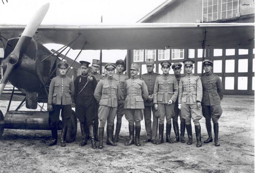 Zwart-witfoto. Groepsfoto van elf militairen op een rij, zij dragen allemaal uniformen met petten en leren laarzen. Sommigen hebben op hun kraag drie sterren, de meesten één. Links een Fokker-vliegtuig, in de achtergrond een hangar.