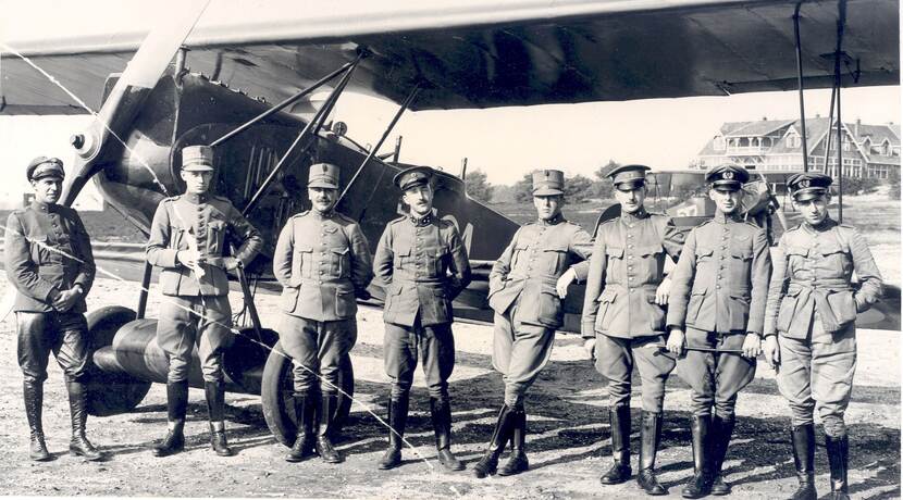 Zwart-witfoto. Groepsfoto van acht militairen op een rij voor een vliegtuig met vleugels boven en onder, zij dragen allemaal uniformen met petten en leren laarzen. De vier rechts leunen op de onderste vleugel van het vliegtuig, de anderen staan met de handen op de rug of voor zich. In de achtergrond een landhuis.