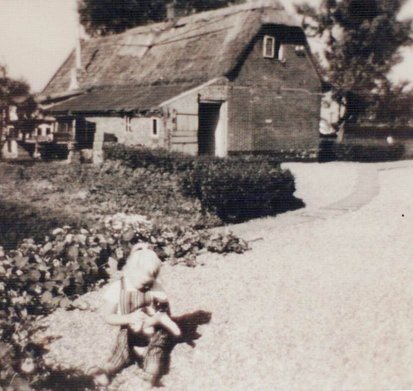 Zwart-witfoto. Een jong meisje zit op de grond in een tuin met een kat in de armen, achter haar een grote tuin met struiken en daarachter een huis met rieten dak. Rechts een brede oprijlaan van grind.