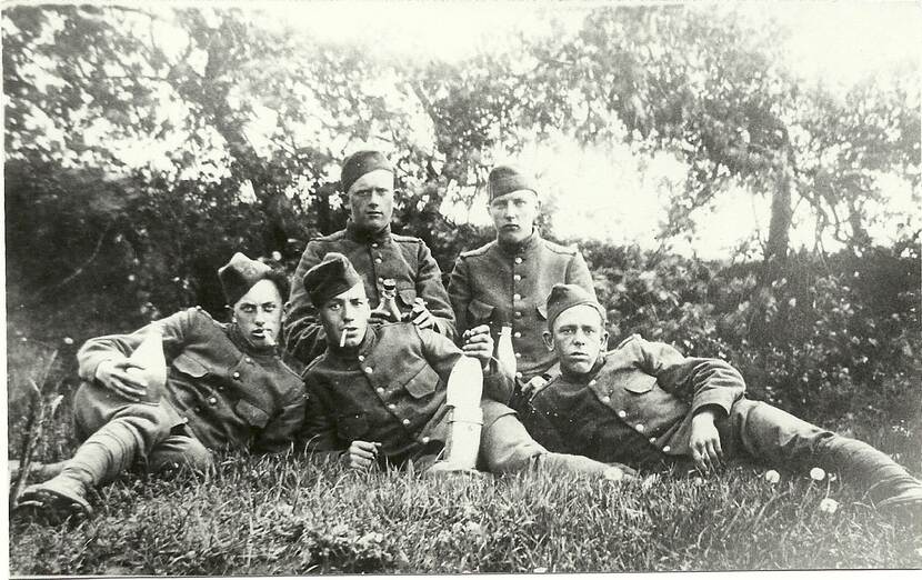 Zwart-witfoto. Vijf militairen in het gras, de voorste drie liggen, de achterste twee zitten, vier houden een fles vast, vier houden een sigaret tussen de vingers of in de mond. Achter hen een heg en bomen.