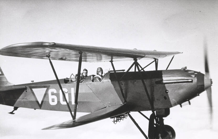 Zwart-witfoto. Een vliegtuig met dubbele vleugels vliegt door de lucht, er zitten twee personen in, ze dragen allebei een vliegermuts en veiligheidsbrillen. Op de zijkant van het toestel is een omgekeerde driehoek en het getal 601 geschilderd.