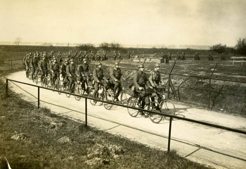 Zwart-witfoto. Een grote groepen militairen fietst over een weggetje tussen weilanden door. Aan weerszijden van het weggetje zijn hekken. De militairen dragen uniformen, helmen en geweren op de rug. Zij fietsen twee-aan-twee.