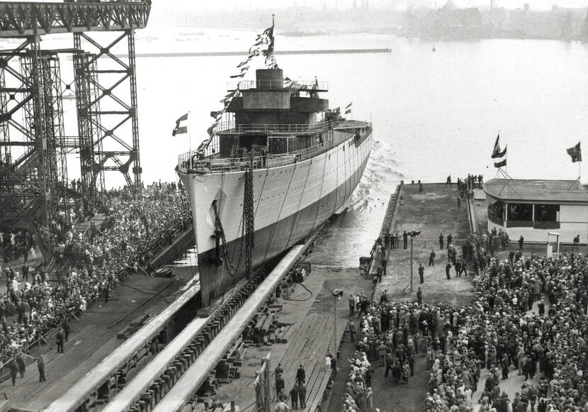 Zwart-witfoto. Een marineschip ligt op een rails en wordt achterstevoren het water in gebracht. Vanaf de punt loopt een lijn met vlaggetjes tot de vlaggenmast in het midden en verder door naar de achterkant. Op de punt is ook een vlaggenmast met de Nederlandse vlag te zien. Links en rechts van het schip staan heel veel mensen op de kade toe te kijken.