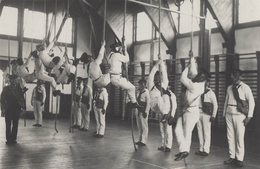 Zwart-witfoto. Tien matrozen, allen in wit gekleed, hangen in een gymzaal aan de touwen. Rechts staat een rij andere matrozen in de houding, links staat een instructeur, hij is in het zwart gekleed. Iedereen, matrozen en instructeur, draagt een gasmasker.