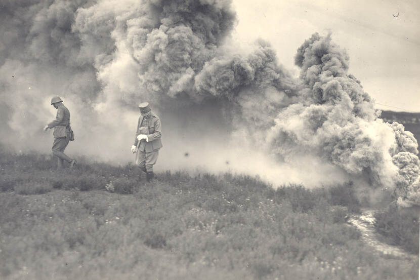 Zwart-witfoto. Twee militairen lopen in een heidelandschap, de linker draagt een helm, de rechter een pet. Achter hen is een enorme rookpluim die het zicht op de achtergrond ontneemt, de militairen draaien hun hoofd en lopen ervandaan.