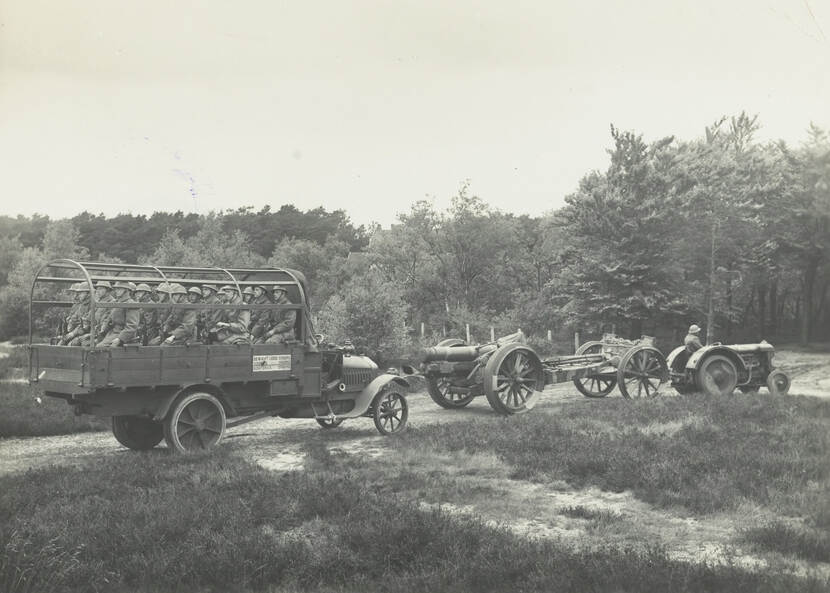 Zwart-witfoto. Links een open vrachtwagen, in de laadbak zitten zestien militairen met hun rug naar de rijrichting. Daarvoor rijdt een houwitsers die wordt voortgetrokken door een tractorachtig voertuig. In de achtergrond bomen en bos.