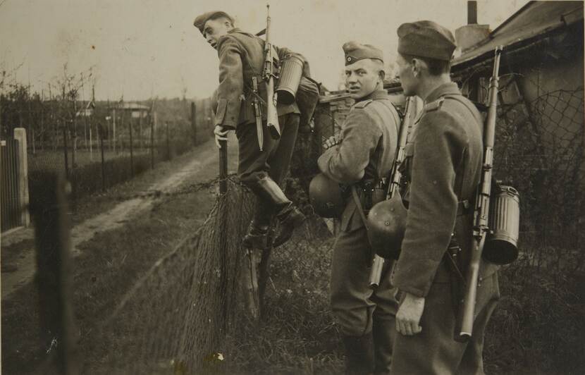 Zwart-witfoto. Drie militairen staan bij een hekje met prikkeldraad, de linker staat op het punt eroverheen te stappen, de middelste kijkt geërgerd achterom naar de camera, de rechter naar het hek.