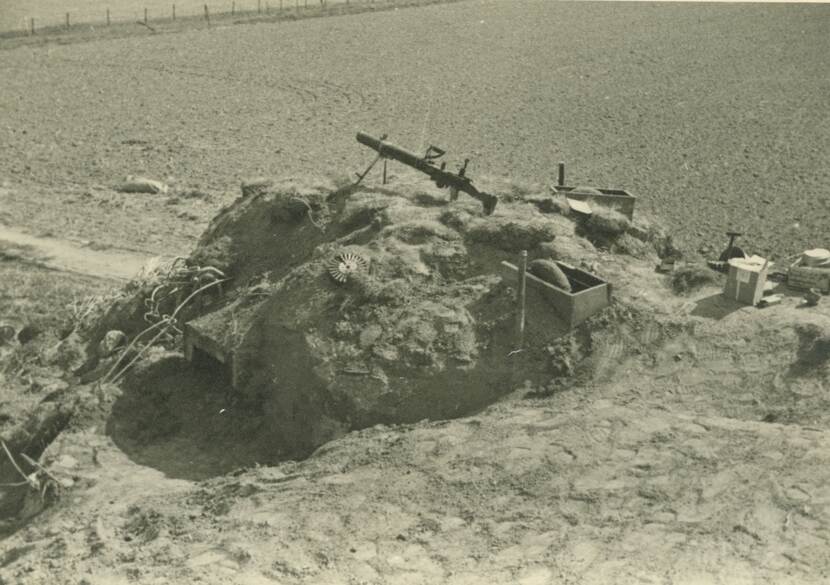 Zwart-witfoto. Een zandhoop met bovenop een mitrailleur. Daaromheen steken twee houten kisten boven het zand uit, rechts wat kartonnen dozen. Links van de mitrailleur ligt een rond magazijn in het zand. In de achtergrond akkerland.