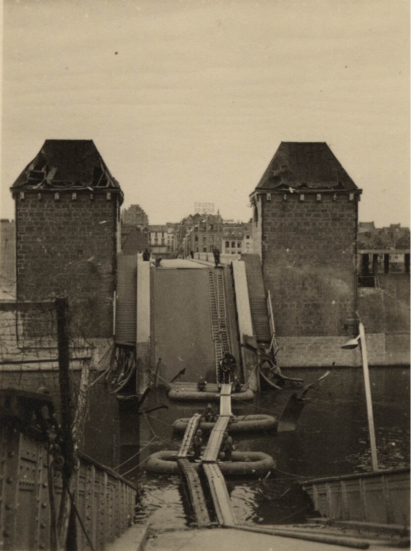 Zwart-witfoto. Uitzicht over een vernielde brug, in het water liggen drie rubberboten met daaroverheen loopplanken die de ene kant met de andere verbinden. Aan de overkant steken aan weerzijde van de weg twee toren uit. Op de rubberen vlotten en bovenaan, tussen de torens, staan een paar militairen. In de achtergrond huizen.
