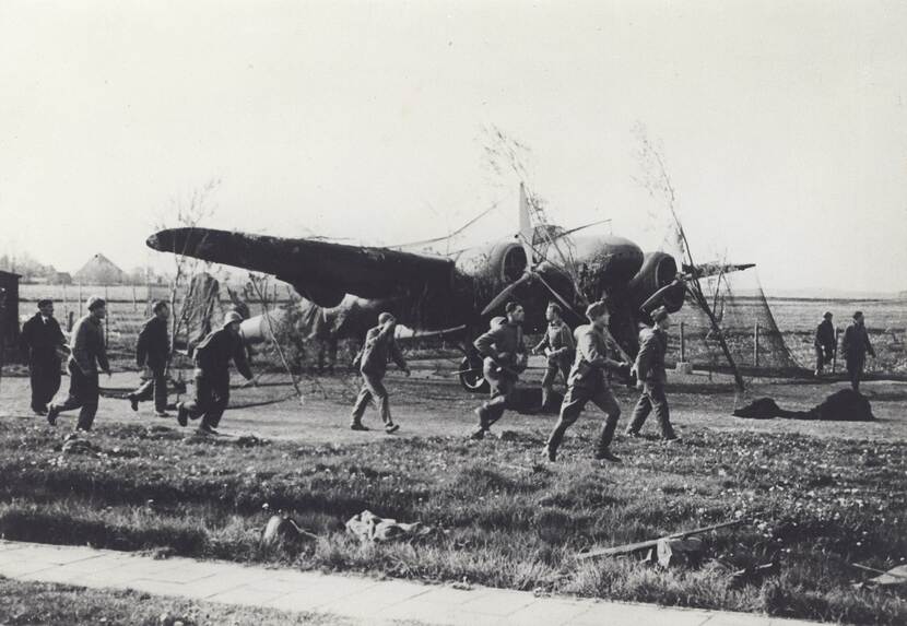 Zwart-witfoto. Een vliegtuig staat op de grond en is met takken en netten gecamoufleerd, daaromheen rennen en lopen militairen, sommigen kijken angstig naar iets rechts buiten beeld.