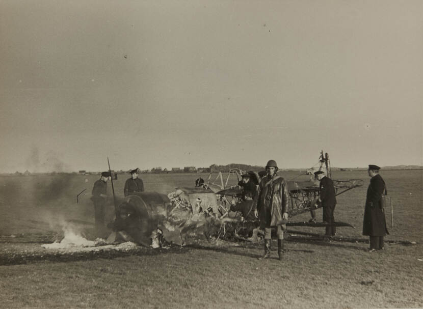 Zwart-witfoto. Het wrak van een vliegtuig ligt in een weiland en staat nog in brand. De piloot staat ernaast, gekleed in lange leren jas en een helm op, hij kijkt in de camera. Zes anderen, politieagenten, inspecteren het brandende restant van het toestel.