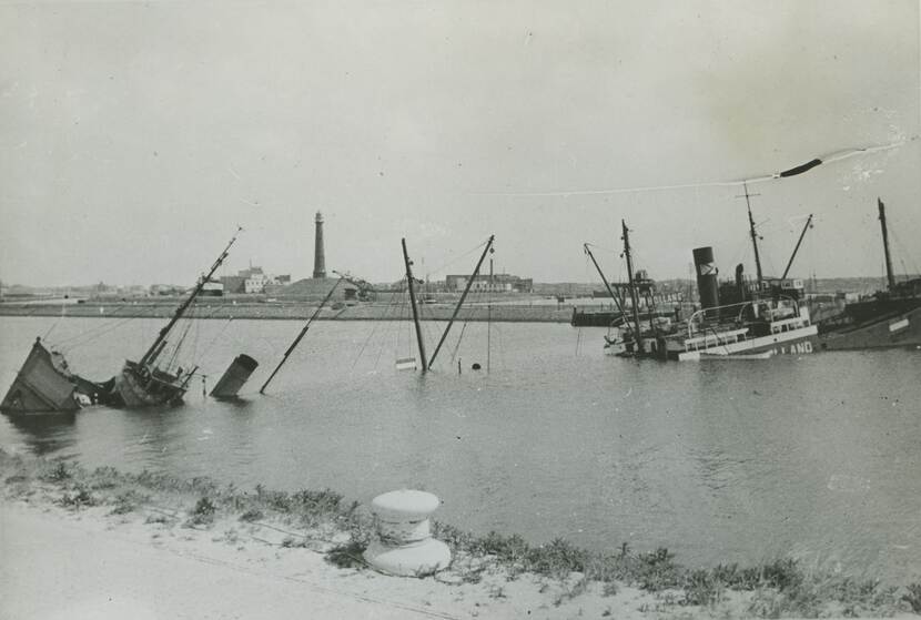 Zwart-witfoto. Een kanaal waar twee schepen in elkaars verlengde tot zinken zijn gebracht. Van de linker steekt alleen de voorpunt, schoorsteen en een paar masten boven het water uit. Van de rechter de achterste helft. In de achtergrond een vuurtoren en havengebouwen.
