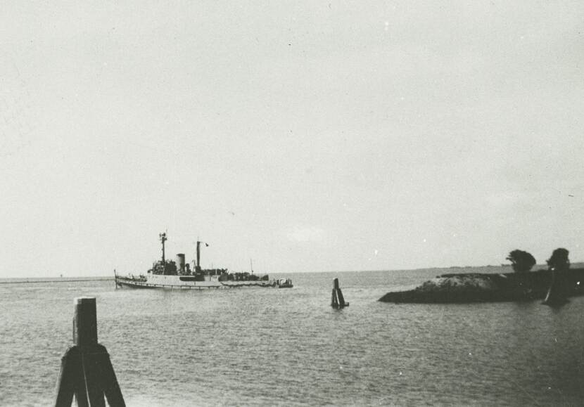 Zwart-witfoto. Een marineschip vaart de zee op. In de voorgrond aanmeerpalen in het water, rechts het uiteinde van een landtong met enkele bomen, het water is rimpelig.