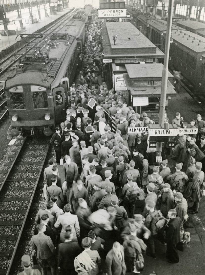 Zwart-witfoto. Van bovenaf genomen, uitkijkend over een perron op een station. Links en rechts van het perron staan treinen klaar, op het perron staan honderden militairen zich te verdringen om in de linker trein te stappen. Vooraan is een paal met bordjes, op de linker staat ‘Alkmaar’ op de rechter ‘Enkhuizen’.