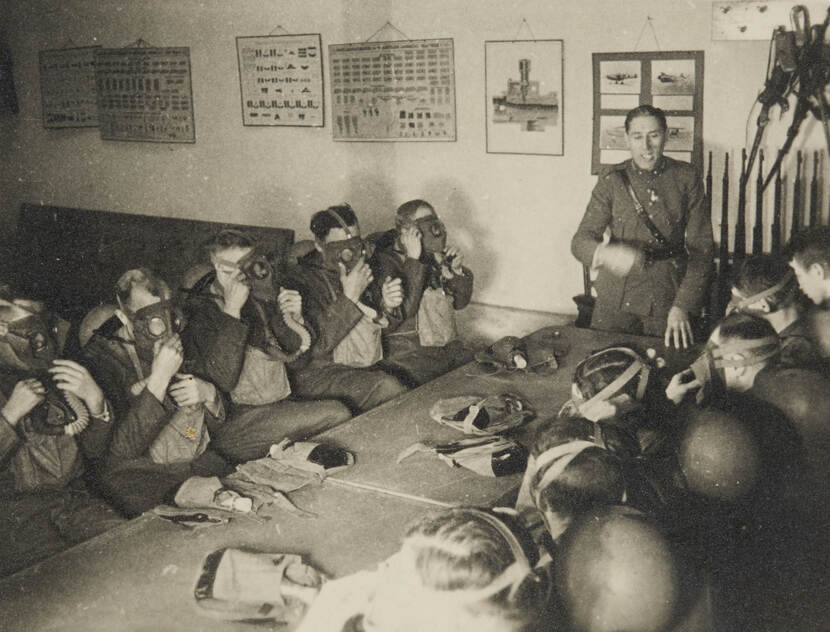 Zwart-witfoto. Elf militairen zitten in vol ornaat aan tafel, hun helmen hangen aan de riempjes aan hun nek, iedereen heeft een gasmasker op. Aan het hoofd van de tafel staat een militair die instructies geeft, aan de muur hangen platen met foto’s, afbeeldingen en schema’s.