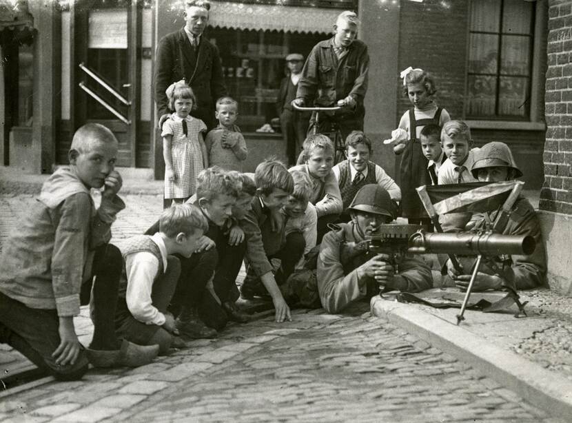 Zwart-witfoto. Straatbeeld, met rechts een militair met mitrailleur die op de grond half op de stoep ligt, naast hem een andere militair. Om hen heen zitten negen jonge kinderen die aandachtig meekijken, daarachter nog drie jongere kinderen, waarvan twee aan de hand van hun vader en een jongen op een fiets. In de achtergrond een winkel.