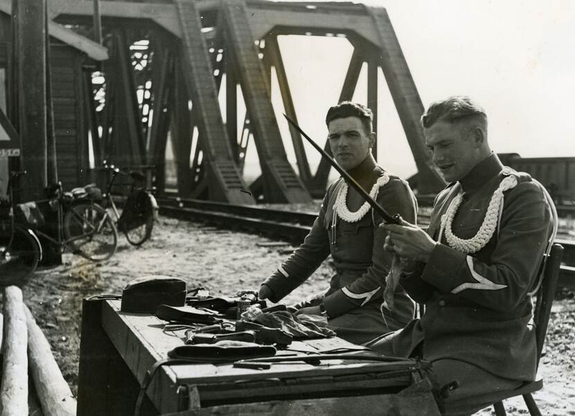 Zwart-witfoto. Twee militairen met een wit koord om hun linkerschouder, zitten voor een tafel waar de onderdelen van hun vuurwapens op liggen. De linker militair kijkt in de camera, de rechter kijkt naar de bajonet en het doekje in zijn handen. Links in de achtergrond twee fietsen, centraal de stalen constructie van een spoorbrug.