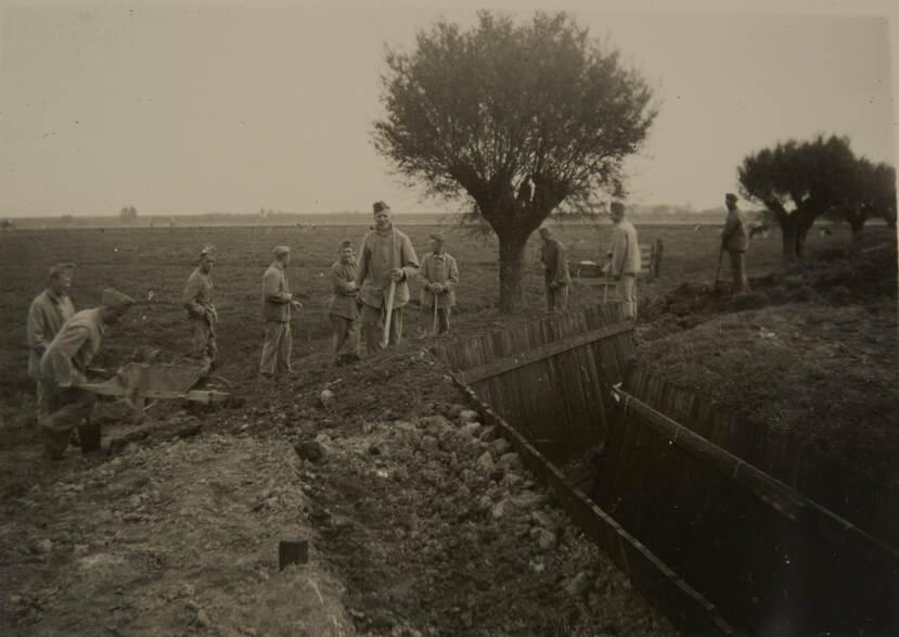 Zwart-witfoto. Een groep van tien militairen staan in een weiland, rechts is een uitgegraven loopgraaf. De militair in het midden rookt een pijp en kijkt naar de camera. Links komen twee militairen met een kruiwagen aanlopen, rechts staan meerdere wilgen langs de loopgraaf.