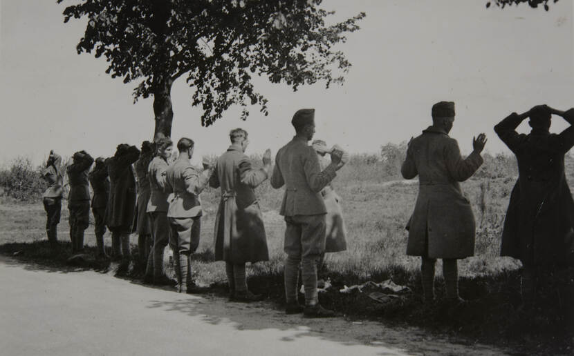 Zwart-witfoto. Twaalf militairen staan langs de kant van de weg, half in de schaduw van bomen, allemaal met de rug naar de camera. Zij geven zich over: velen houden beide handen omhoog, een enkele houdt zijn handen op het achterhoofd. In de achtergrond bermen met hoog gras, struiken en bomen.