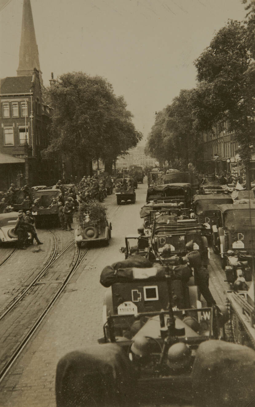 Zwart-witfoto. Straatbeeld, links en rechts staan tientallen militairen vrachtwagens en andere voertuigen langs de kant van de weg geparkeerd. In elke wagen zitten meerdere militairen of staan meerdere militairen ernaast.