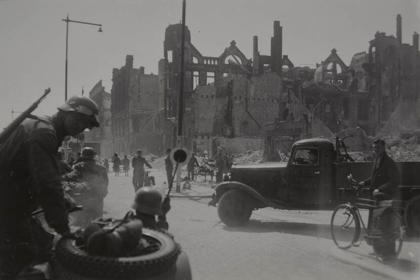 Zwart-witfoto. Links een militair met Duitse helm en een geweer op zijn rug die in de camera kijkt, rechts iets verder weg een burger met fiets aan de hand die ook in de camera kijkt. Om hen heen meerdere militairen en burgers die op straat lopen, rechts draait een vrachtwagen de straat op. Veel burgers hebben een fiets aan de hand. In de achtergrond puin en ruïnes van kapotgeschoten gebouwen.