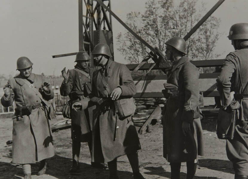 Zwart-witfoto. Vier Nederlandse militairen, gekleed in lange jassen en helmen op, staan voor een hoge ijzeren constructie, zij houden hun handen omhoog in overgave. Rechts staat een Duitse militair (rug naar de camera) met zijn handen in de zij naar hen te kijken.