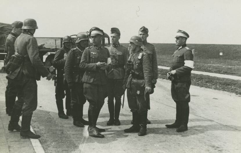 Zwart-witfoto. Een groep van negen militairen staat langs de kant van de weg te overleggen. Sommigen dragen een witte band om hun arm en hoofddeksel. In de achtergrond een militair voertuig en duinlandschap.