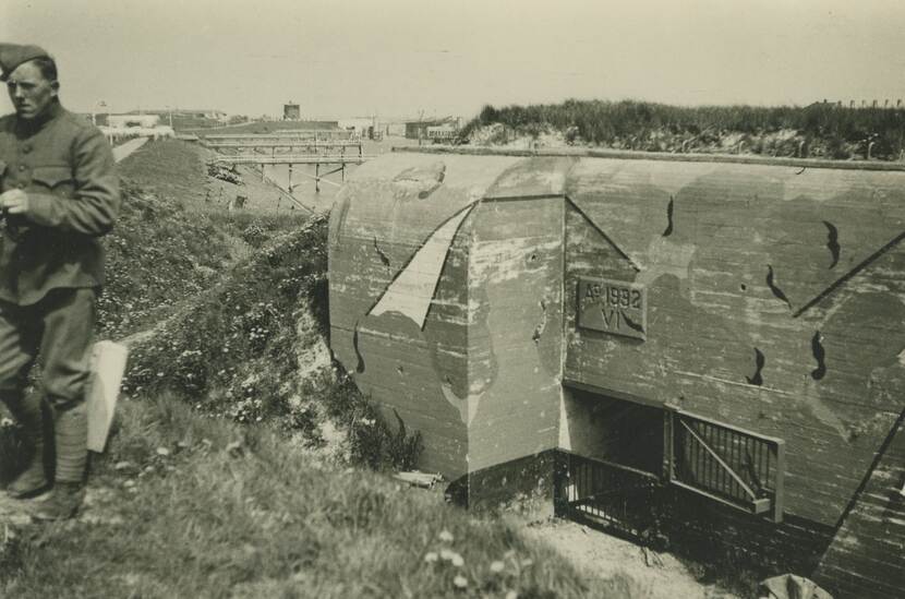 Zwart-witfoto. Een bunker in een duinlandschap, boven de ingang staat ‘Ao 1992’ met daaronder ‘VI’, het Romeinse cijfer zes. Op de betonnen buitenkant zijn een paar camouflageschilderingen aangebracht. Helemaal links staat een Nederlandse militair bovenop de duin, in de achtergrond aanlegsteigers in een kleine zeearm.