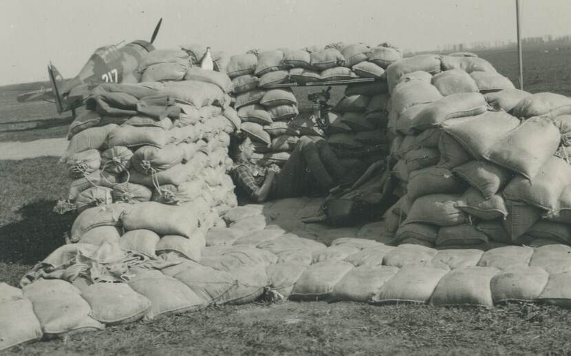 Zwart-witfoto. De achterkant van een manshoge verdedigingsopstelling opgebouwd uit zandzakken, centraal daarin een mitrailleur die door een opening in de zandzakken naar buiten staat gericht. In de opstelling ligt een militair te slapen. Links in de achtergrond een vliegtuig met het cijfer 217 op de zijkant geschilderd.