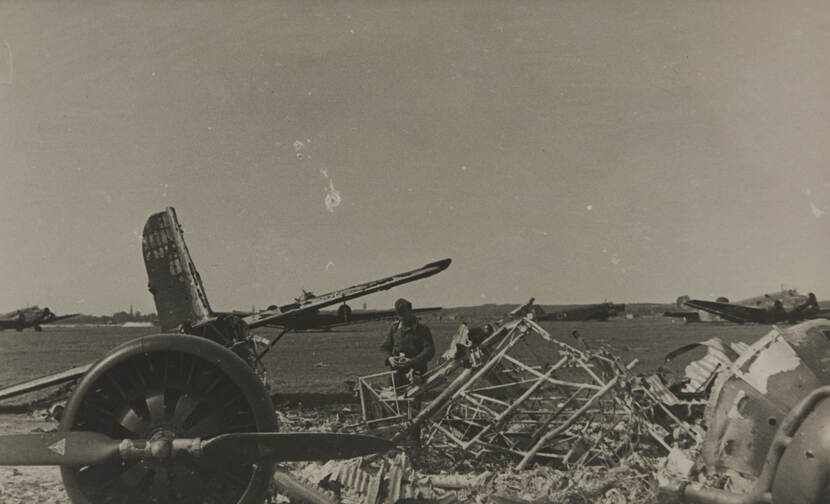 Zwart-witfoto. Een militair staat tussen het puin van een neergestort en uitgebrand vliegtuig. Links ligt een grote motor met propeller, daarachter is de staart van het vliegtuig te zien. De militair houdt een camera vast om een foto te maken. In de achtergrond staan meerdere vliegtuigen aan de grond.