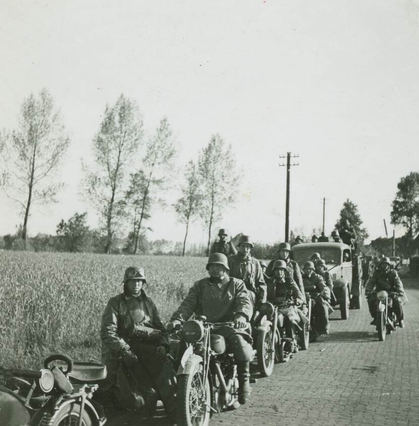 Zwart-witfoto. Een colonne van vijf militaire motoren (met zijspan) en een vrachtwagen staan op een landweggetje, op en om de voertuigen kijken ongeveer vijftien militairen naar de camera. Rechts komt een motor aanrijden. In de achtergrond een akker, bomen en elektriciteitskabels.