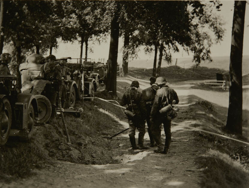 Zwart-witfoto. Een weg over een dijk, aan beide kanten geflankeerd door bomen. Links een rij geparkeerde militaire voertuigen, op de weg zijn drie militairen met iets aan het zeulen. Aan de linkerkant van de weg is een vers gedolven graf met een pet erop.
